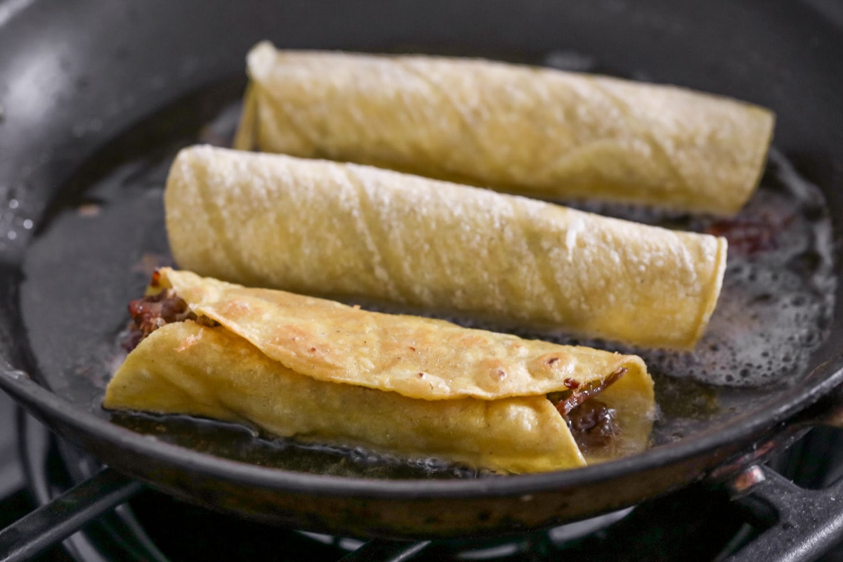 Flautas being fried in a frying pan with a little bit of oil