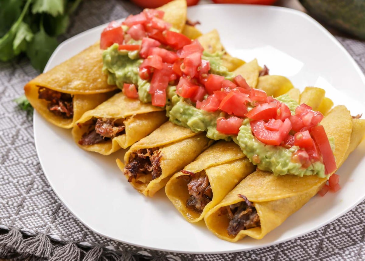 Beef Flautas topped with guacamole and tomatoes on a white plate