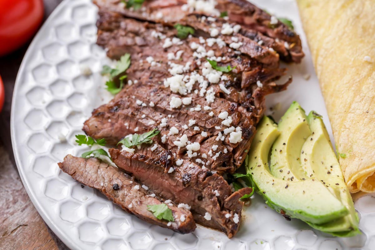 Carne asada cut in strips with sliced avocado and rolled tortillas on the side on a white plate.