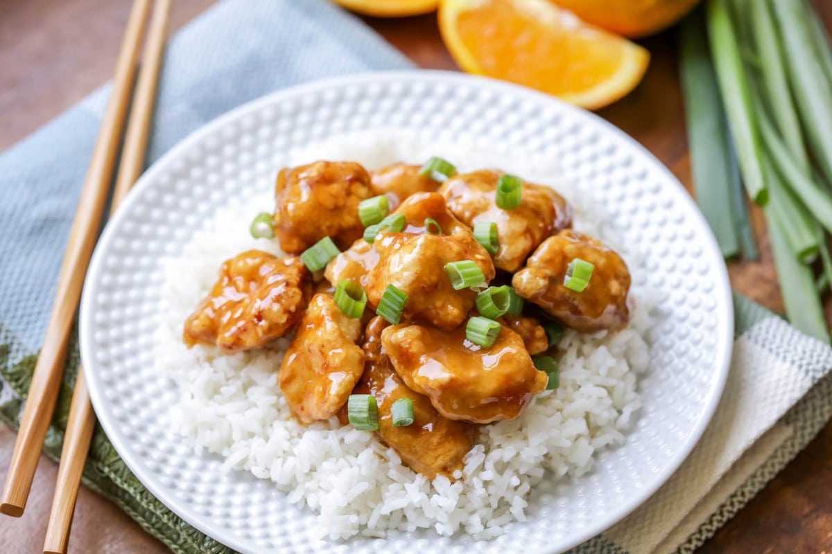 Asian Dinner Recipes - Orange Chicken garnished with chopped green onions on white rice on a white plate. 