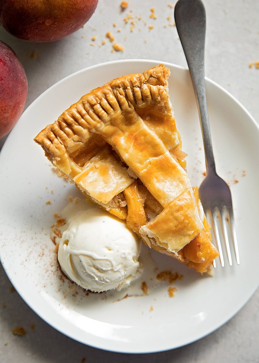 Slice of fresh peach pie recipe served on a white plate with vanilla ice cream.