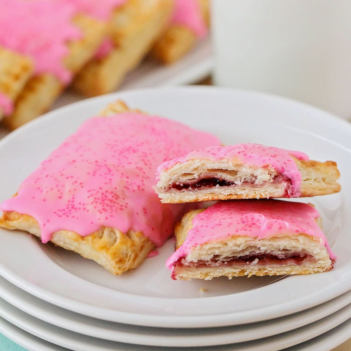 Homemade pop tarts with filled with fruit preserves and topped with a pink frosting with pink sugar crystals on a stack of white plates. 