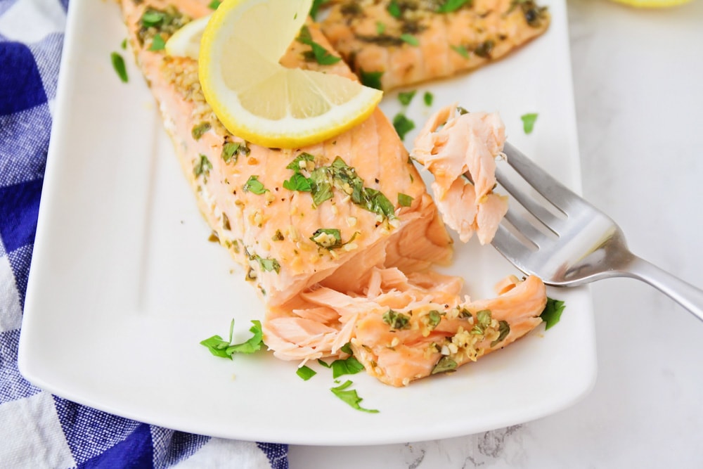 Baked Salmon on a serving platter garnished with chopped parsley and a lemon slice with a metal fork resting on the platter. 