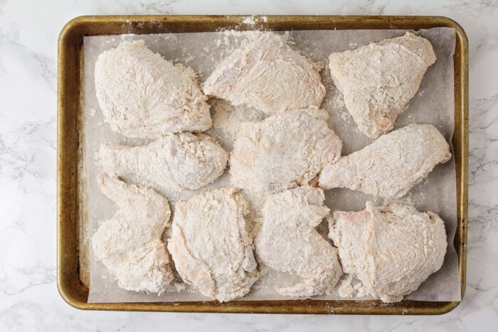 Chicken dipped in buttermilk and flour on a lined baking sheet.