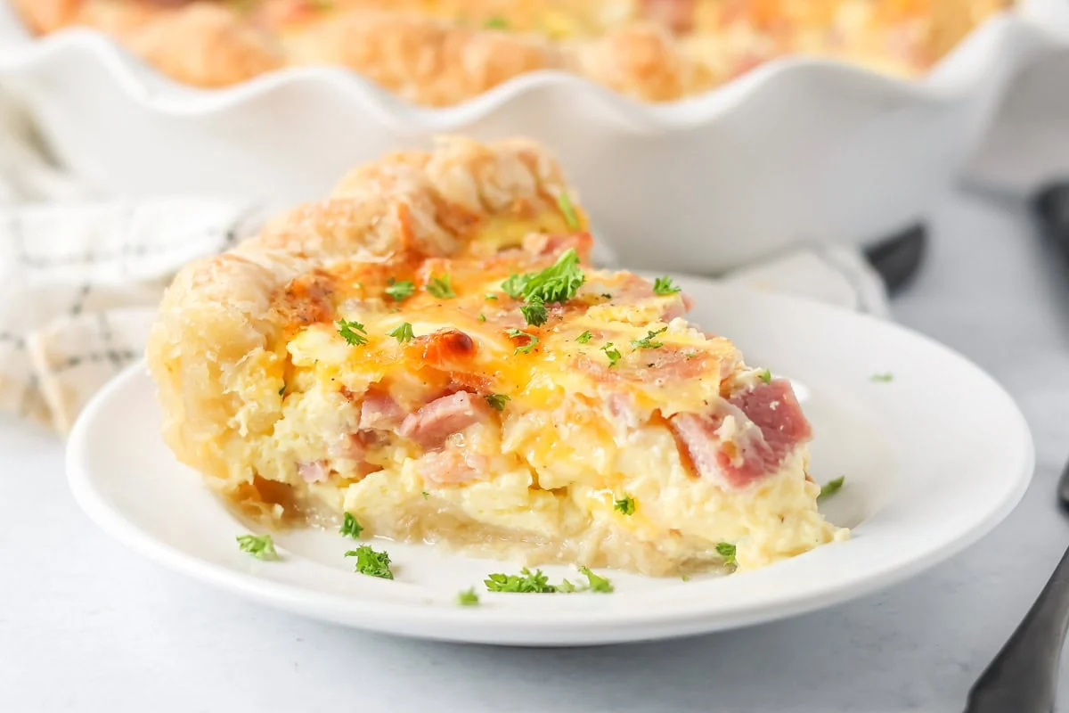 Slice of ham and cheese quiche recipe on white plate.