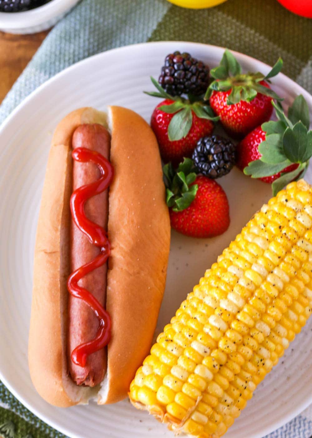 Boiled hot dog in a bun on a plate with corn and fruit on the side