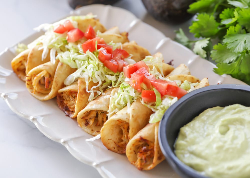 Flautas on a white platter with a small bowl of dip on the side.