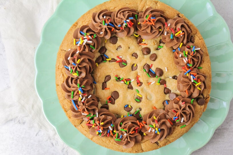 Celebrate any occasion with a chocolate chip cookie cake recipe topped with chocolate buttercream frosting and colorful sprinkles! Homemade Chocolate chip cookie cake recipe topped with frosting and sprinkles.