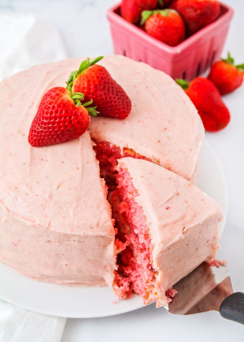 Strawberry frosting on strawberry cake with two strawberries on top and a slice being cut out of the cake closeup pic.