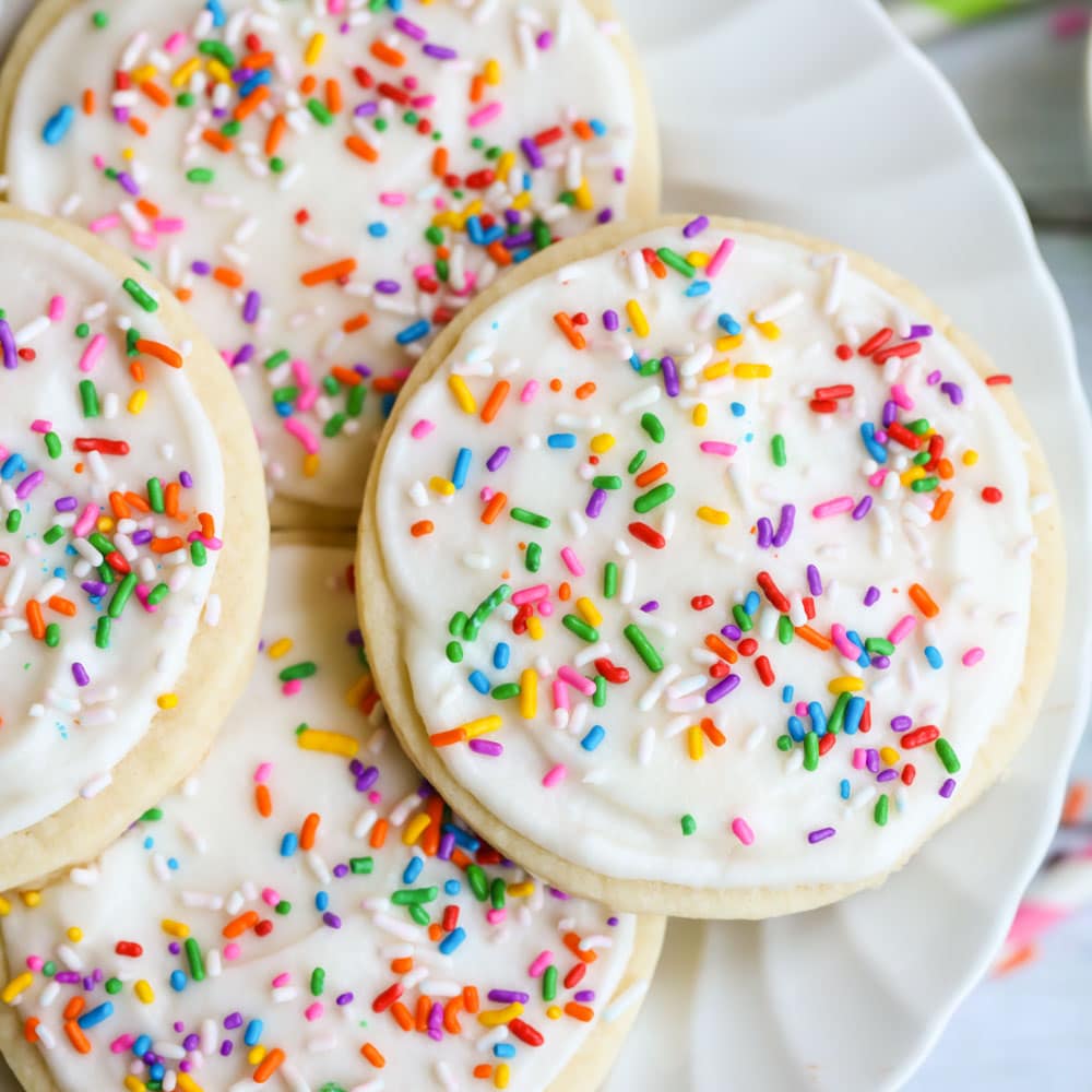 Four sugar cookies on a white plate with sugar cookie frosting and colorful sprinkles. 