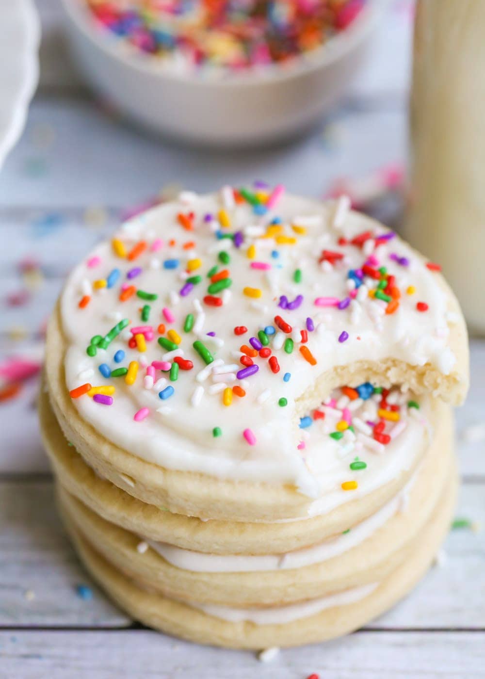A stack of four sugar cookies with sugar cookie icing and colorful sprinkles, with a bite taken out of the top cookie