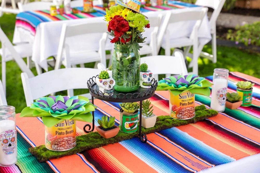 A table outdoors with a Mexican blanket-type covering and various Mexican decorations on top with a similarly decorated table in the background.