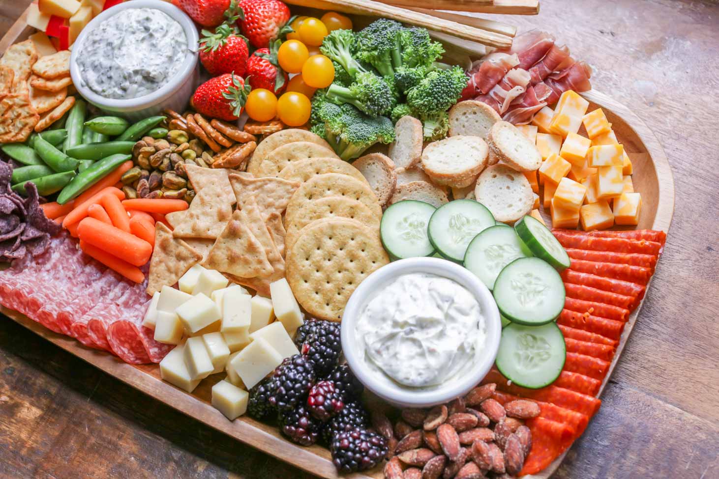 A charcuterie board with crackers, cheese, veggies, and dips.