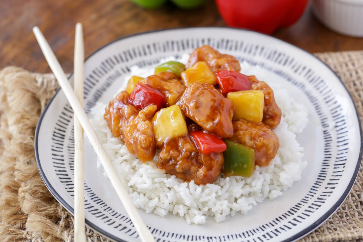 Asian Dinner Recipes - Sweet and sour chicken on top of white rice on a white plate with wooden chopsticks resting on it. 