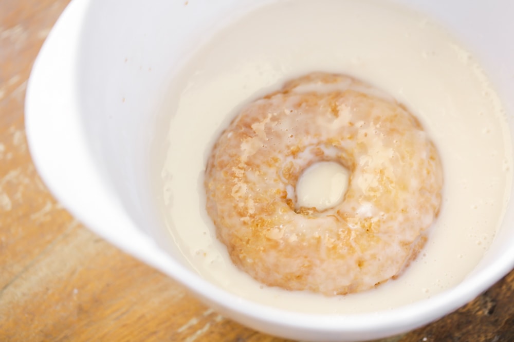 A glazed buttermilk donut being dunked in the bowl of glaze