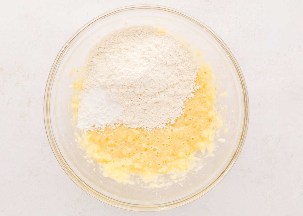 Adding flour to a banana mixture in a glass bowl.