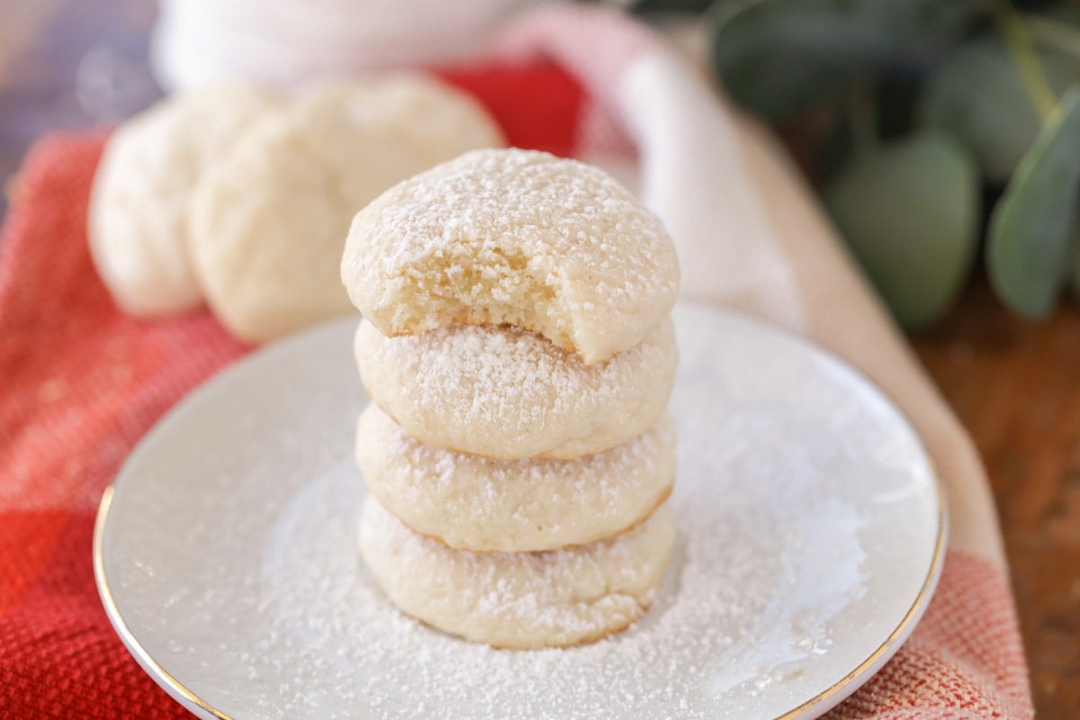 Cream cheese cookies stacked on a white plate, covered in powdered sugar