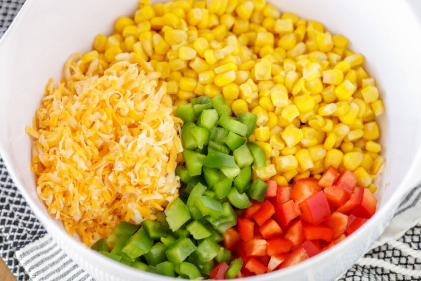 Corn, cheese, and diced bell peppers in a white bowl for frito salad.