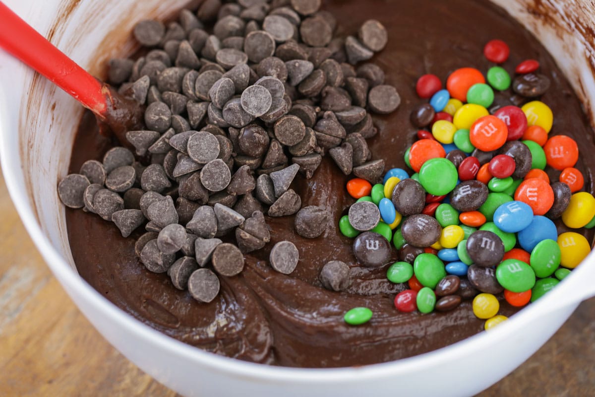 Brownie batter, chocolate chips, and m&ms in a white mixing bowl