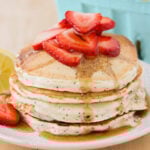 A stack of Lemon Poppy Seed Pancakes topped with sliced strawberries and syrup.