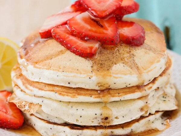 A stack of Lemon Poppy Seed Pancakes topped with sliced strawberries and syrup.