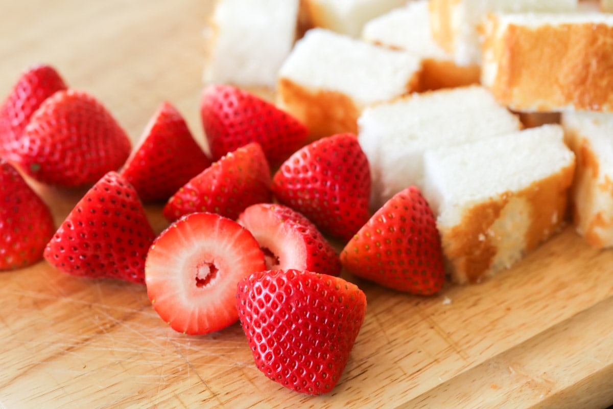 Sliced strawberries and cubes of white cake on a wood cutting board.