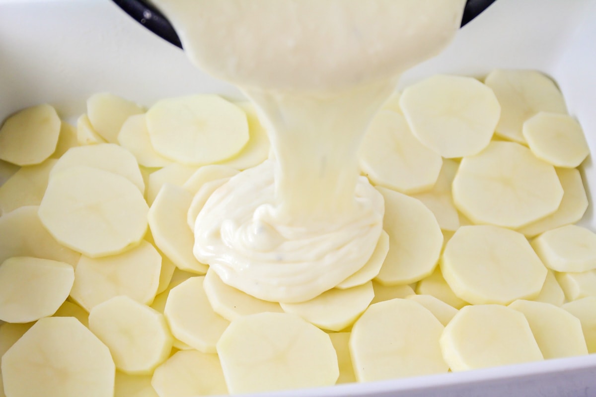 Pouring cream sauce over potato slices in a white casserole dish