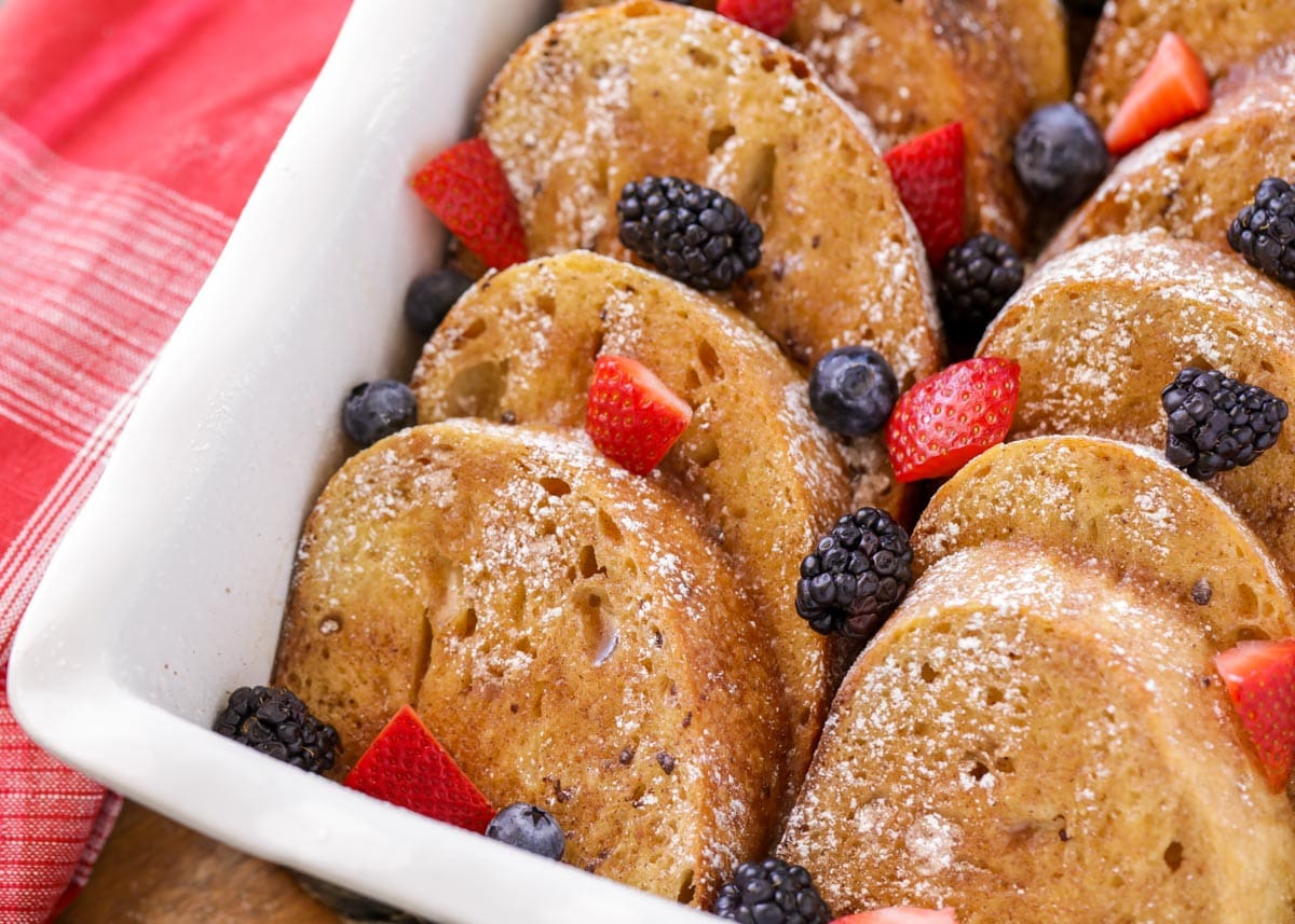 Oven French Toast sprinkled with powdered sugar and topped with fresh berries in a white baking dish. 