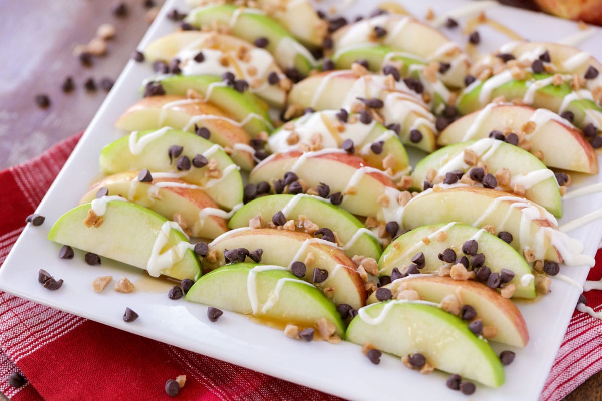 Sliced red and green apples on a white platter topped with caramel sauce, a white chocolate drizzle, mini chocolate chips, and toffee bits. 