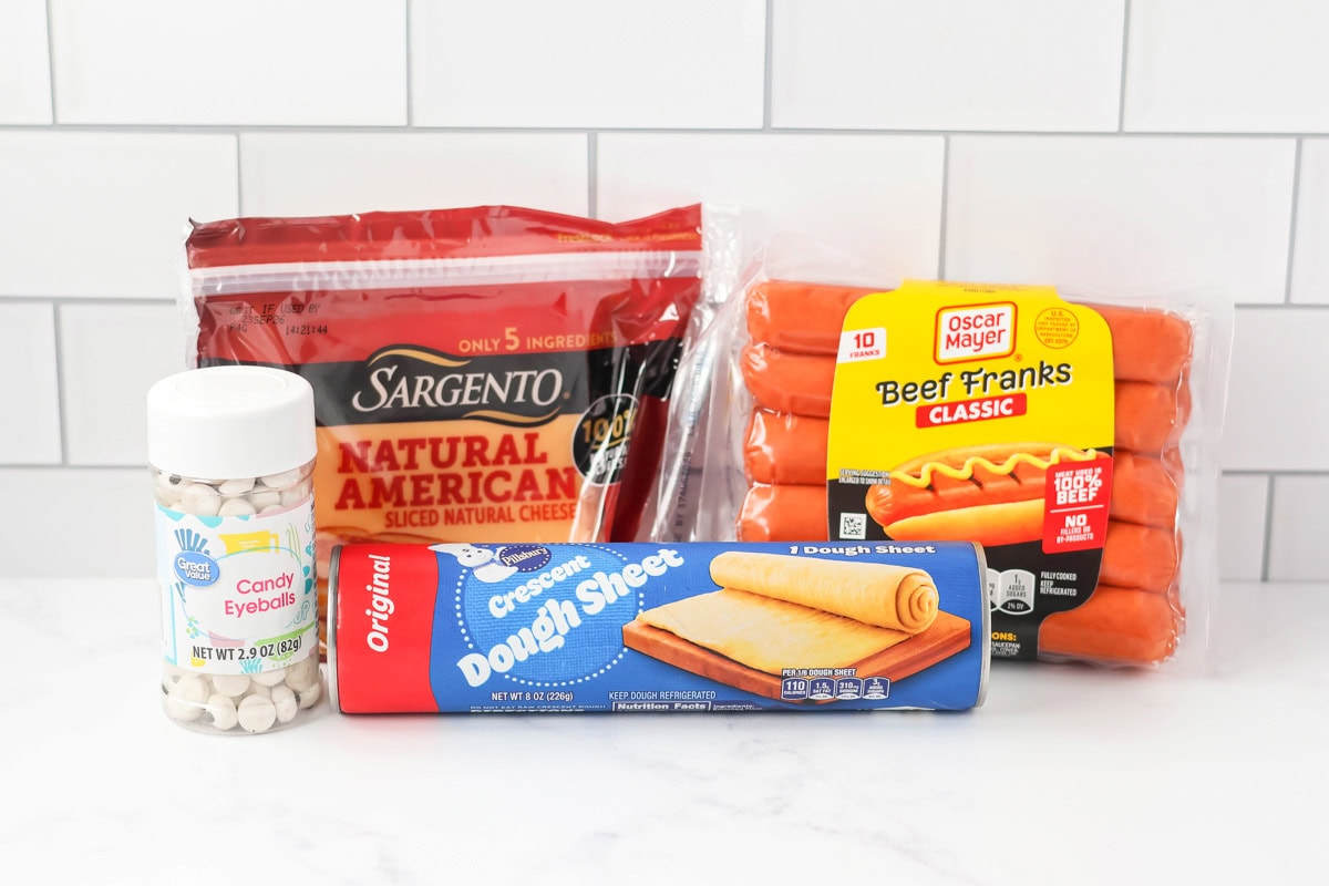Buttery crescent-wrapped mummy hot dogs are filled with cheese and topped with cute candy eyes for spooky kid-friendly fun!! Mummy hot dogs ingredients on counter.