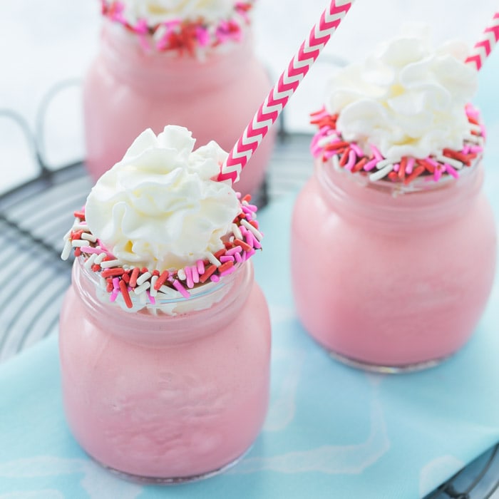 Valentine's Day Desserts - Red Velvet Milkshakes in small Mason jars with sprinkles on the rims topped with whipped cream and pink and white chevron patterned straws. 