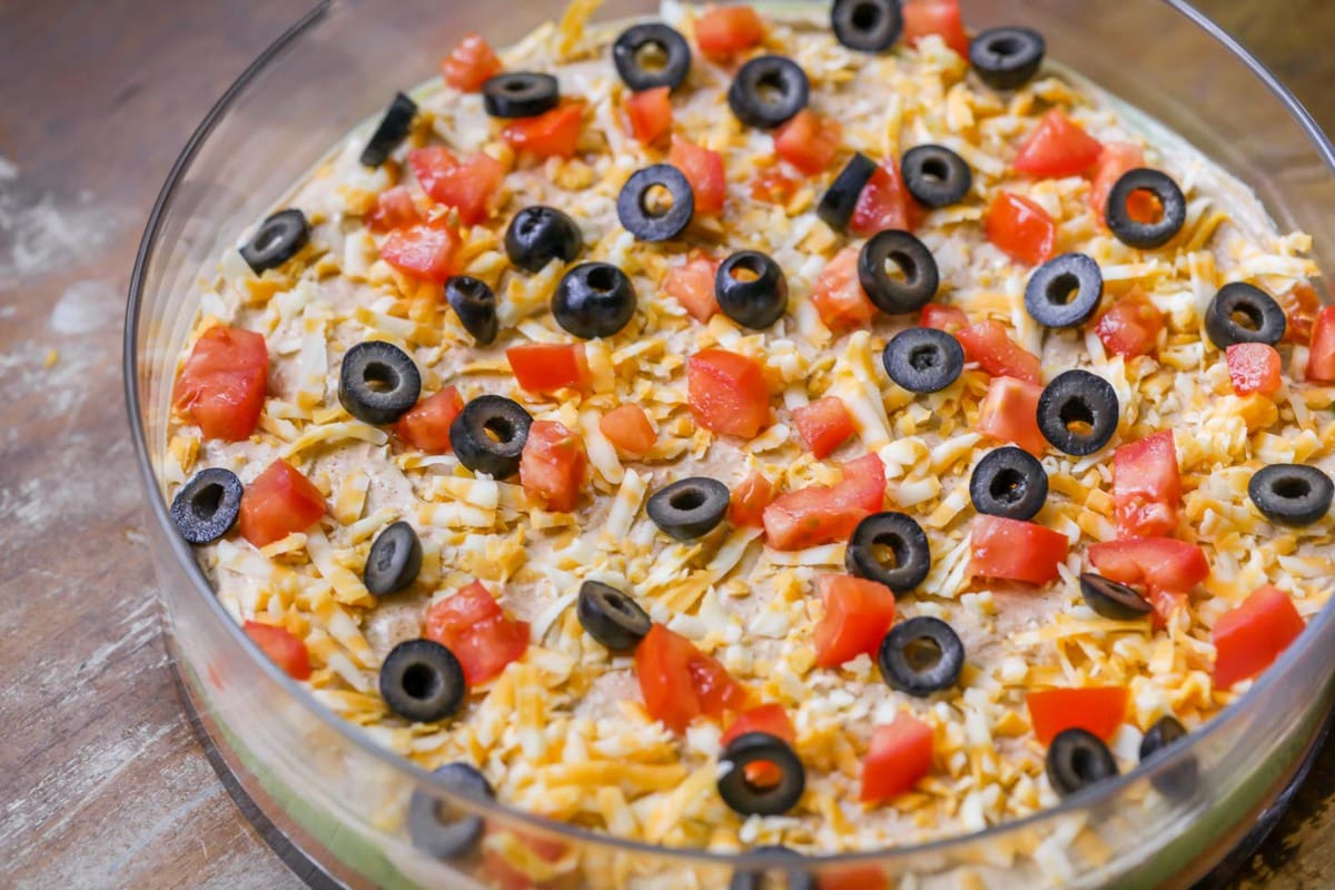 Super Bowl Appetizers - 7 Layer Bean Dip in a clear glass bowl topped with sliced black olives and diced tomatoes. 