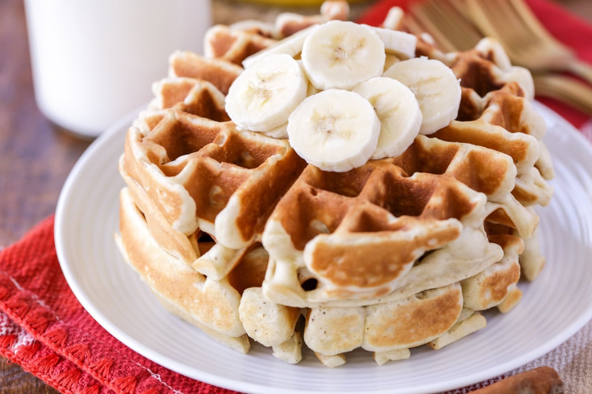 Two banana waffles stacked on a white plate topped with sliced bananas. 