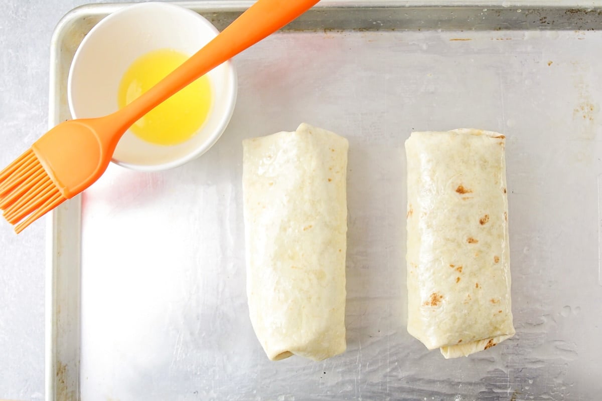 Brushing two wrapped burritos with oil and prepping for baking.