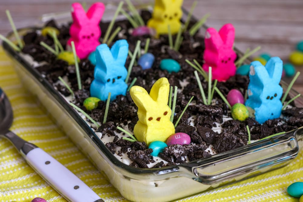 Easter dirt cake topped with crushed Oreo cookies, Easter Bunny Peep marshmallow candies, and egg shaped candies in a glass baking dish. 