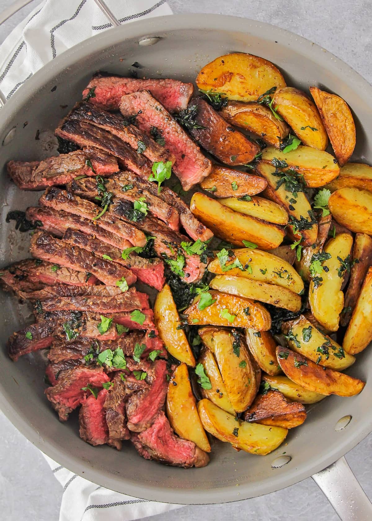 Sliced steak and potato wedges served in a skillet and topped with fresh herbs.