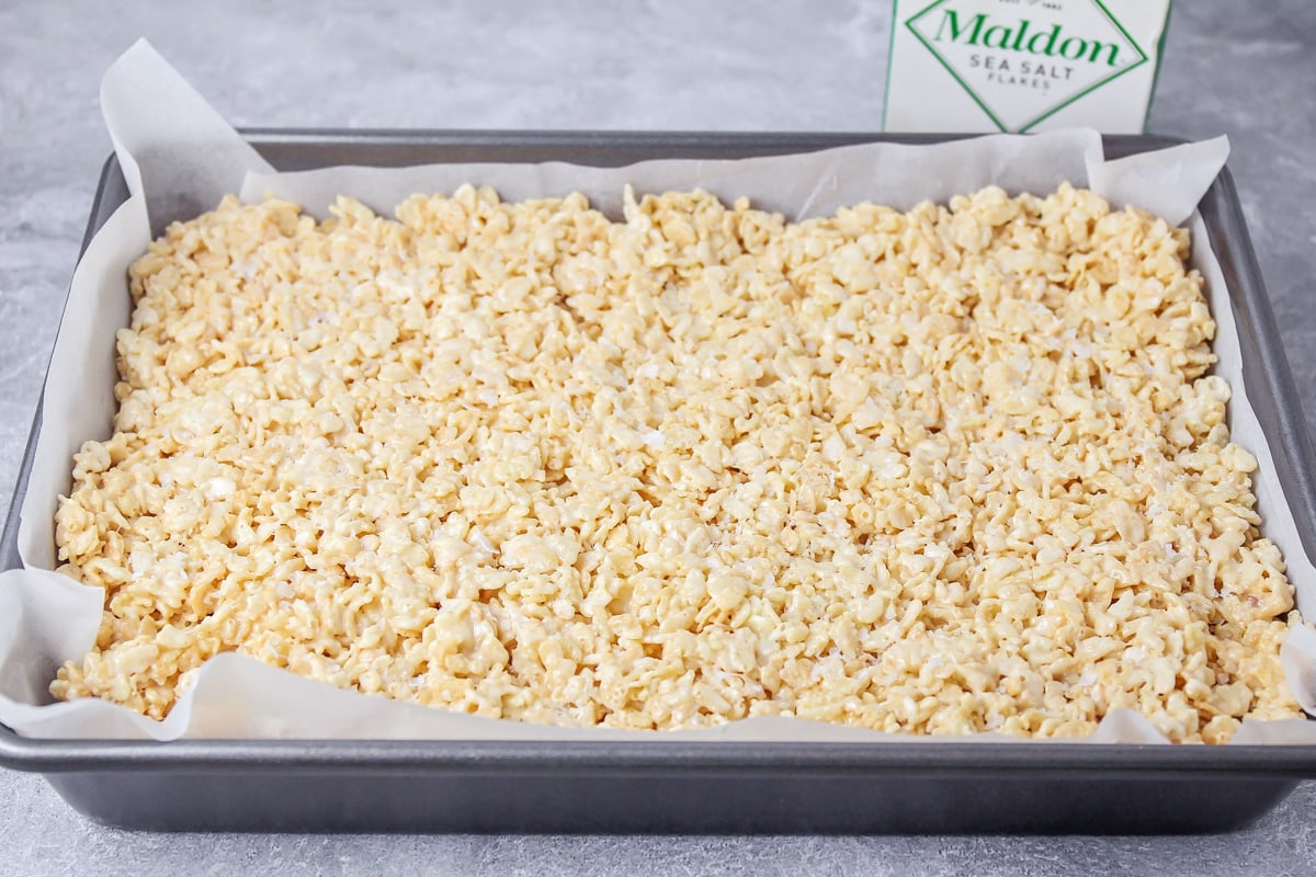 Pressing rice krispie treats into a metal pan lined with parchment paper.
