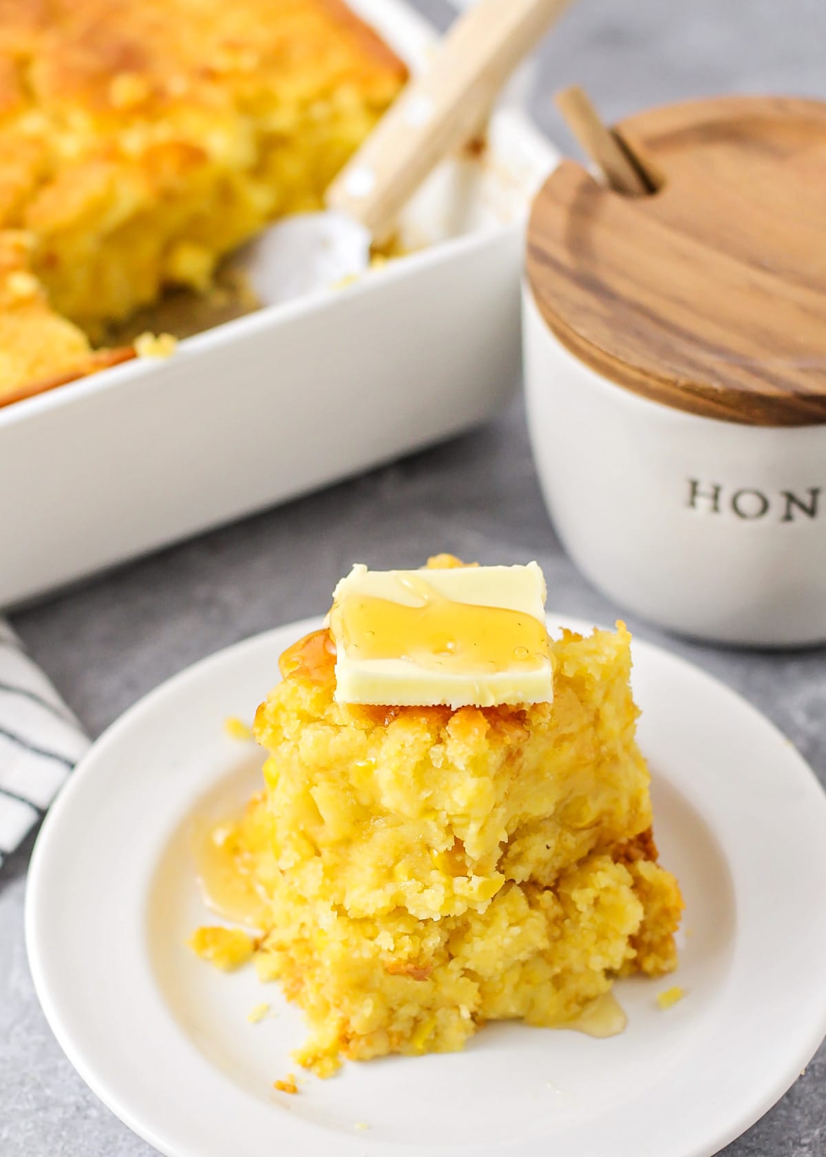 Slices of gooey cornbread casserole stacked on a white plate and topped with butter and honey.