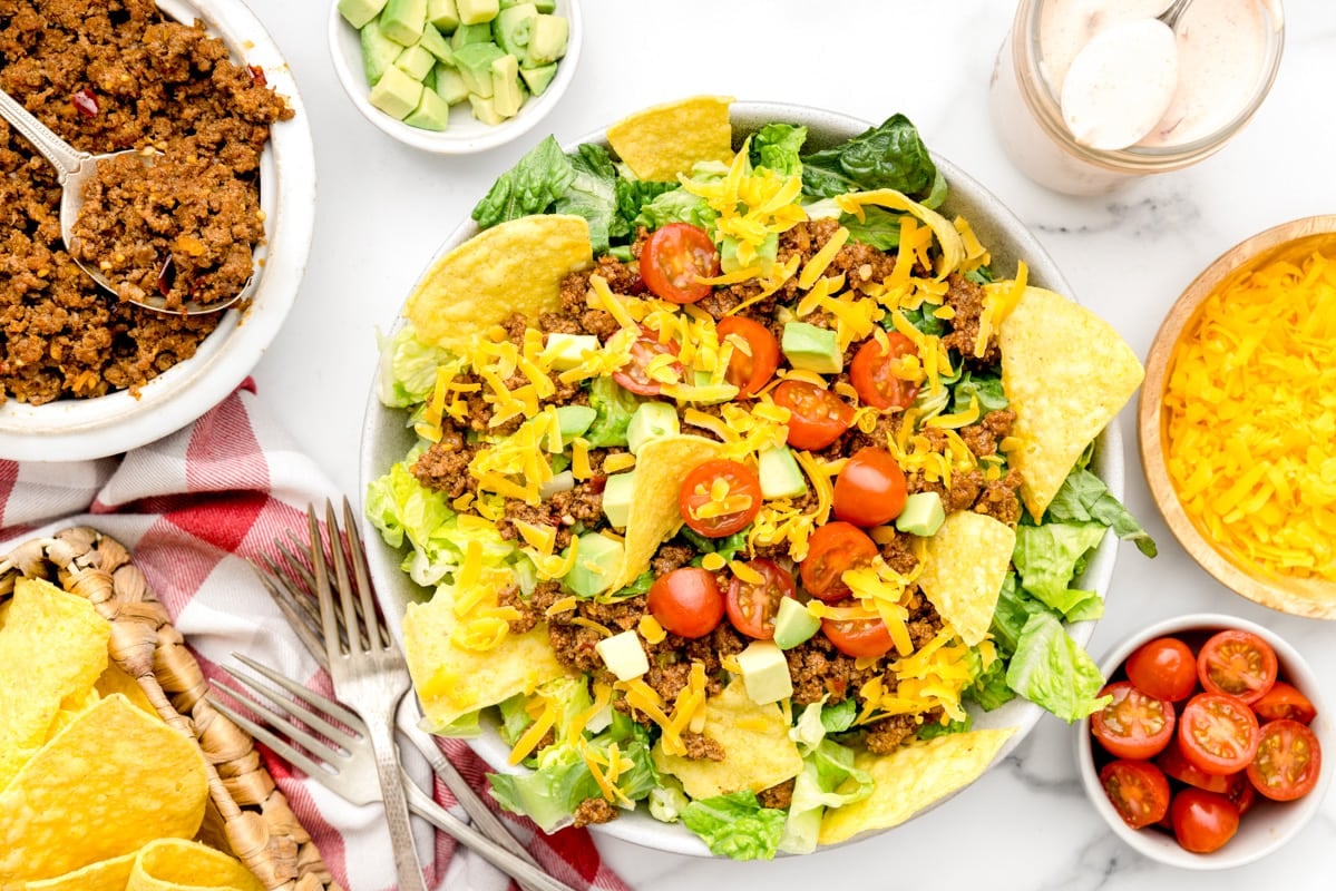 Taco salad topped with diced cherry tomatoes, shredded cheddar cheese, ground beef, diced avocados, and garnished with corn tortilla chips in a white bowl. 
