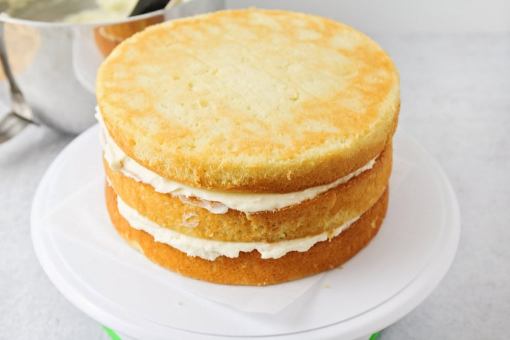 Three layers of homemade vanilla cake being assembled on cake stand.
