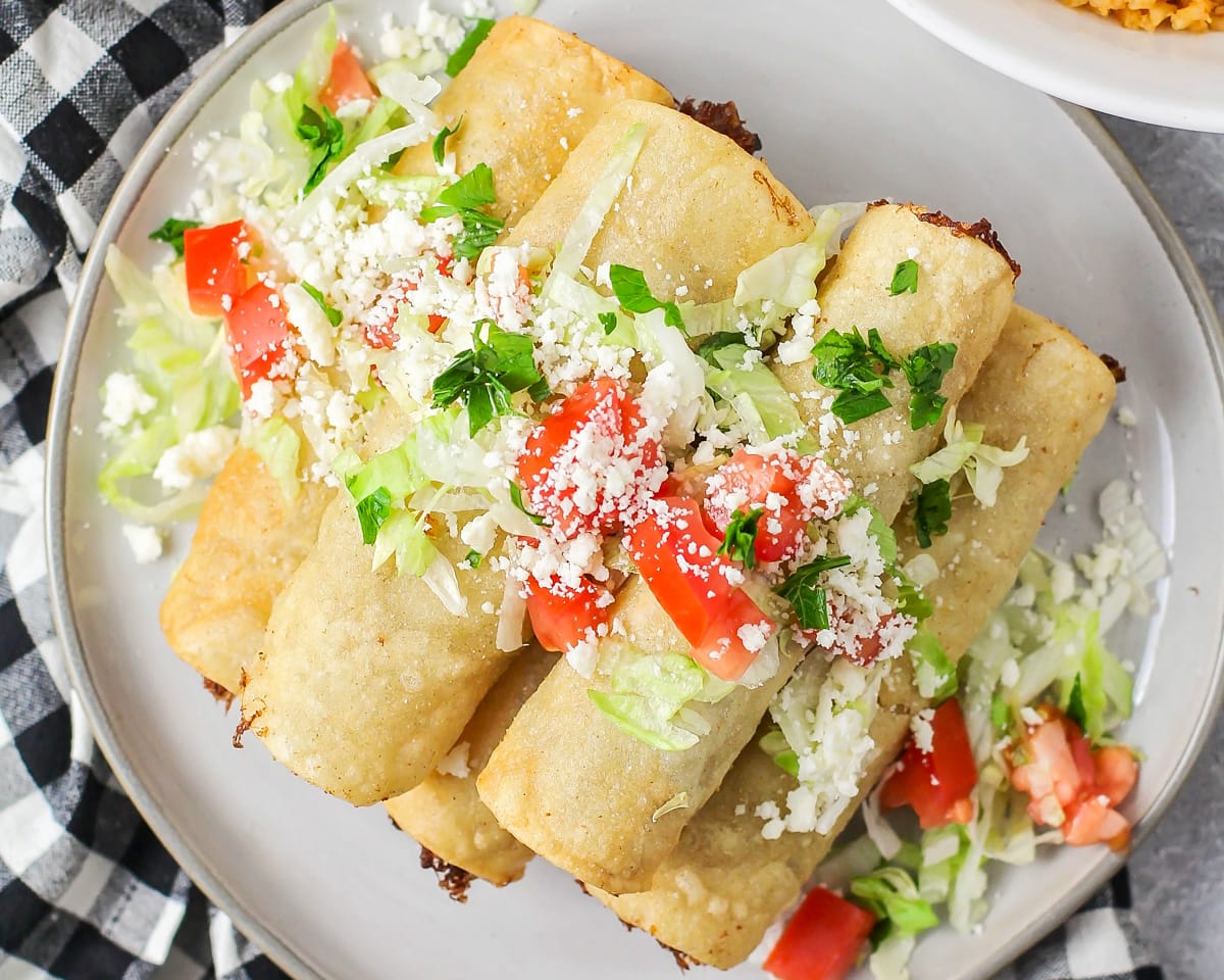 A pile of flautas topped with cotija, tomatoes, and cilantro.