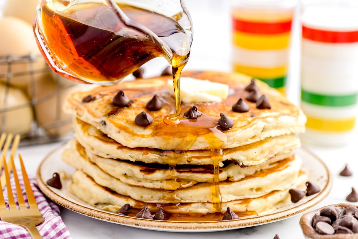Pouring syrup onto a stack of 5 fluffy buttermilk chocolate chip pancakes.