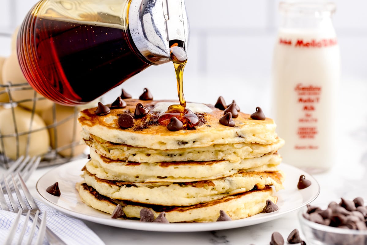 Pouring syrup on a stack of 5 chocolate chip pancakes.