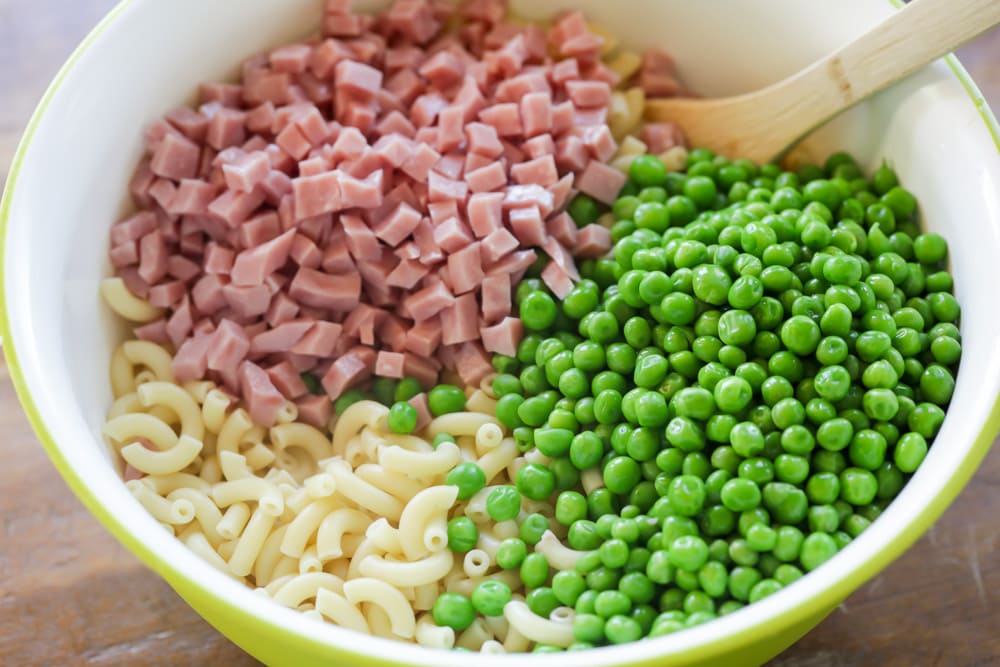Macaroni, ham chunks, and peas in a yellow bowl ready for mixing.