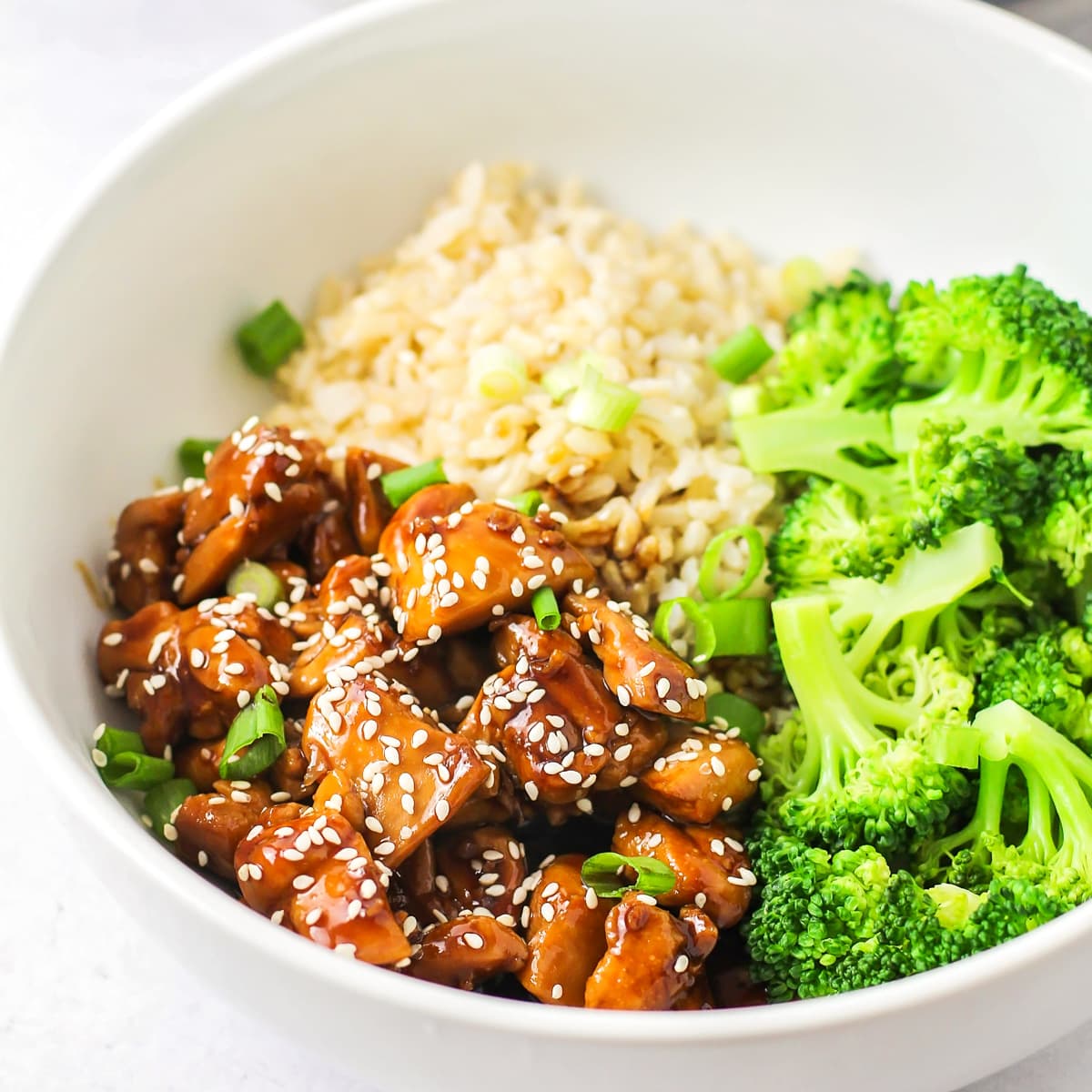 Teriyaki sauce coating chicken in a white bowl served with rice and broccoli.