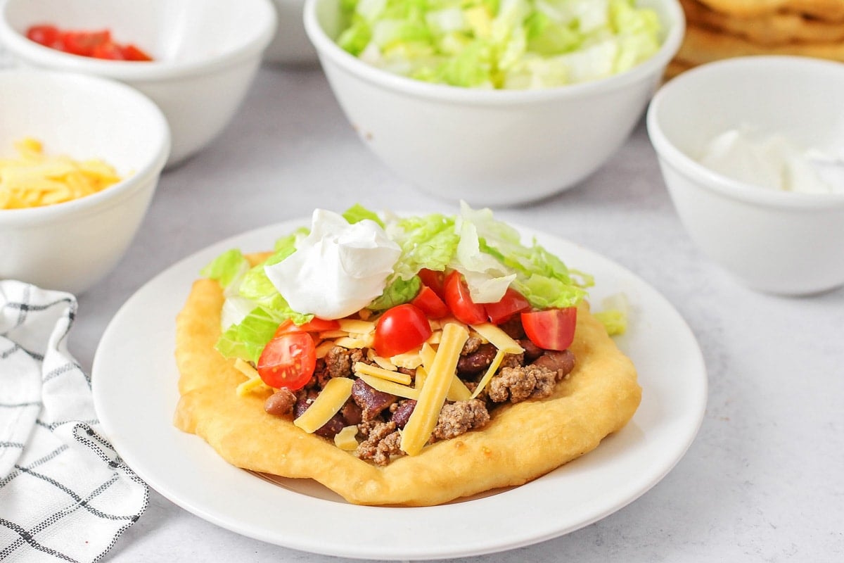 Indian Fry Bread topped with a beef and bean mixture, shredded cheddar cheese, diced tomatoes, lettuce, and a dollop of sour cream on a white plate. 