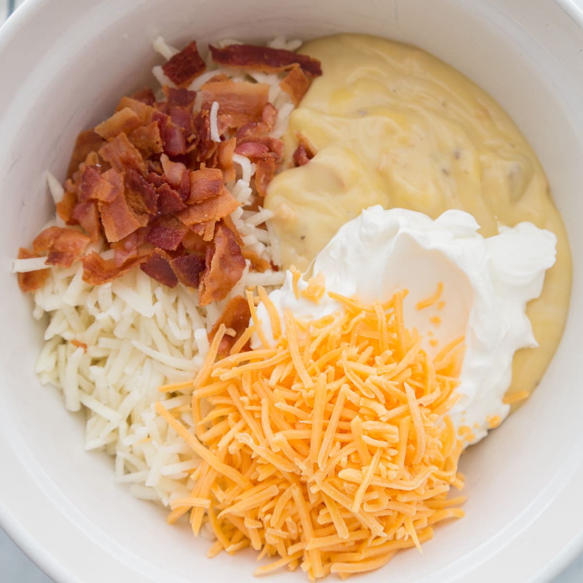 Cream soups, bacon, cheese, and hashbrowns mixed in a white bowl.