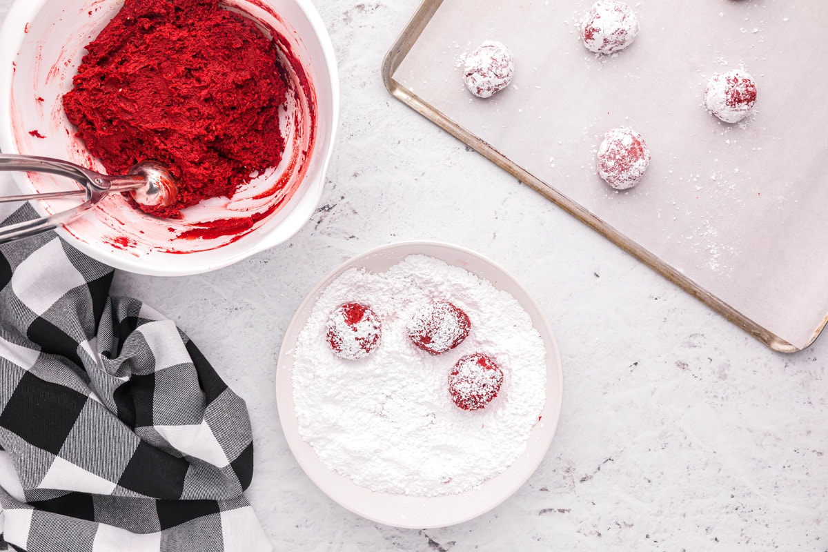 Scooping red velvet cake mix cookies batter and rolling in powdered sugar.