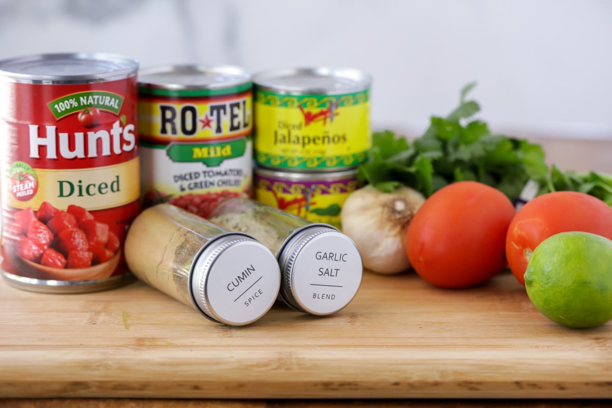 Ingredients for homemade salsa laid out including a can of diced tomatoes, Ro-Tel, cumin, garlic salt, fresh tomatoes, lime, garlic, and cilantro. 