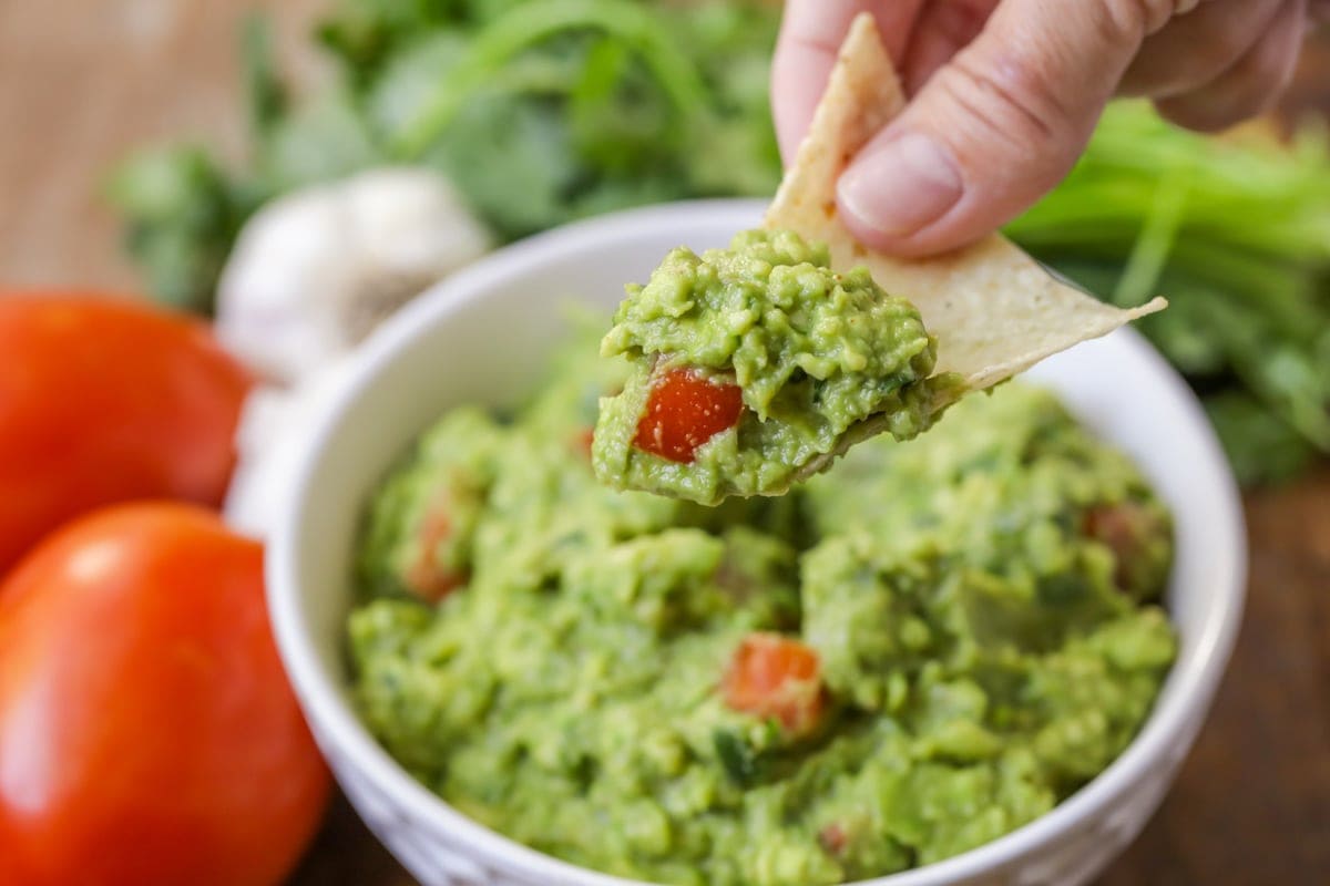 A hand holding a chip with guacamole on it with a bowl of guacamole and ingredients to make it in the background.